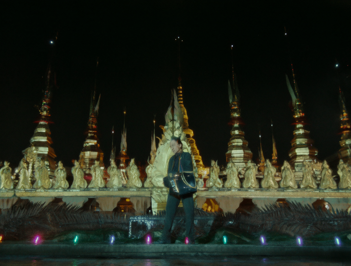Vista cinematográfica de un hombre frente a espléndidas torres doradas bajo un cielo nocturno con reflejos neón, capturando la atmósfera sintética y exótica de Bangkok.