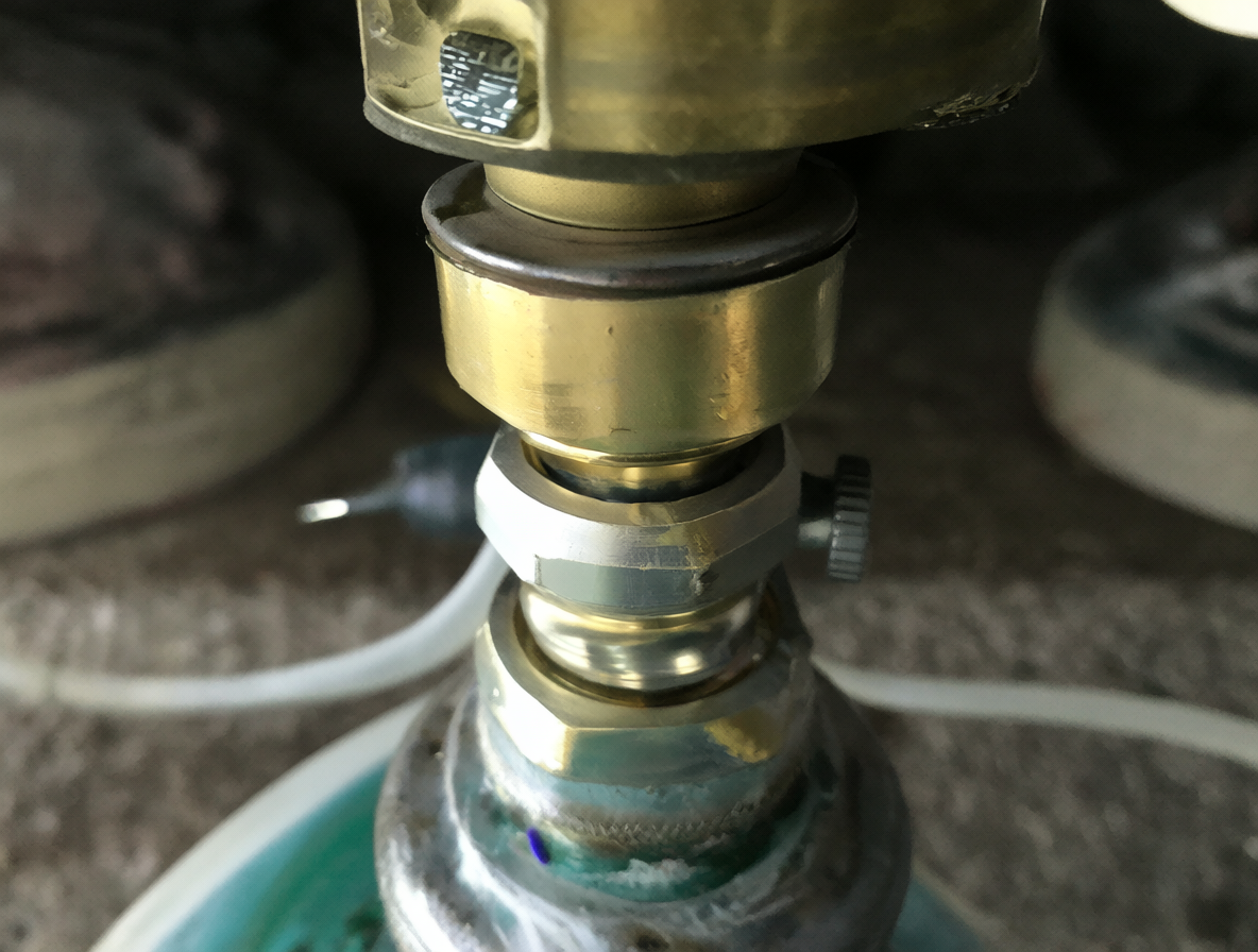 Primer plano técnico del regulador de gas conectado a una manguera flexible con el cilindro al fondo desenfocado.