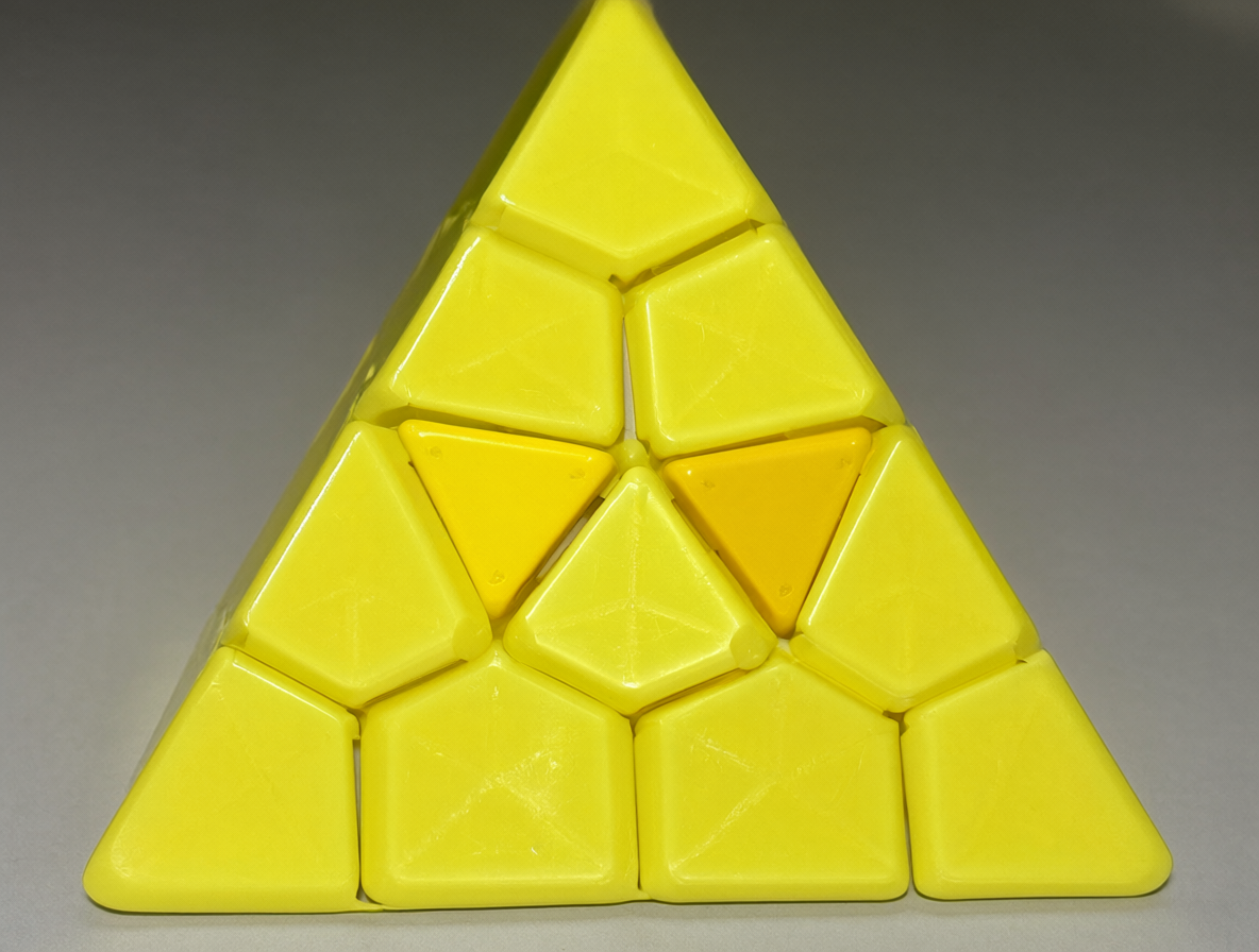 A close-up photo of a yellow plastic Pyraminx puzzle, showing the specific detail of two central face pieces being swapped.