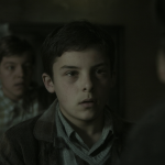 Plano cinematográfico mostrando a un estudiante en la Europa de la posguerra, con una figura borrosa detrás pronunciando sus pensamientos internos.
