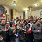 Grupo de personas cantando con banderas chilenas dentro de una iglesia iluminada.