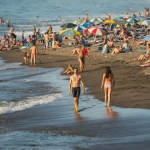 Vista de un hombre y una mujer paseando por una playa oscura llena de gente bajo el sol.
