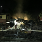 Vagones de carga y pasajeros destrozados sobre las vías principales, humo subiendo al cielo oscuro tras un accidente grave.