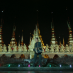 Vista cinematográfica de un hombre frente a espléndidas torres doradas bajo un cielo nocturno con reflejos neón, capturando la atmósfera sintética y exótica de Bangkok.