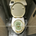 Vista dividida de un baño público mostrando el espacio debajo de una puerta y materiales económicos como madera barata o plástico oxidado.