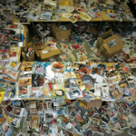 Vista cenital de un espacio desbordado con cientos de revistas en formato A3 amontonadas sobre mesas y estanterías, junto a cajas archivadoras abiertas.