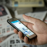 Close-up of a hand refreshing a smartphone screen against a backdrop of Japanese newspaper headlines.