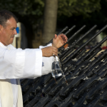 Fotografía de un sacerdote bendiciendo armas de fuego, mostrando el contraste entre la vestimenta religiosa y los rifles modernos.