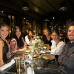 Fotografía de un hombre joven sentado en una mesa con varias mujeres sonrientes en un restaurante elegante.