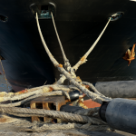 Vista detallada de un gran buque portacontenedores atracado en un muelle industrial, mostrando claramente las líneas de amarre gruesas de acero extendiéndose desde los cabos del barco hacia los bolardos del puerto, con grandes defensas elásticas de goma comprimidas entre el casco y la estructura de hormigón.