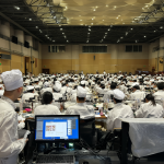 Vista del salón de conferencias en un instituto, lleno de estudiantes usando batas de chef blancas. Al frente, una persona está cerca de una impresora 3D mostrando software en su pantalla.