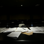 Vista de un salón de audiencias oscuro con una pila de documentos etiquetados como SQM sobre un escritorio.