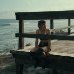Vista panorámica de una chica sentada en una tarima de madera junto al mar, con una baranda y el océano al fondo.